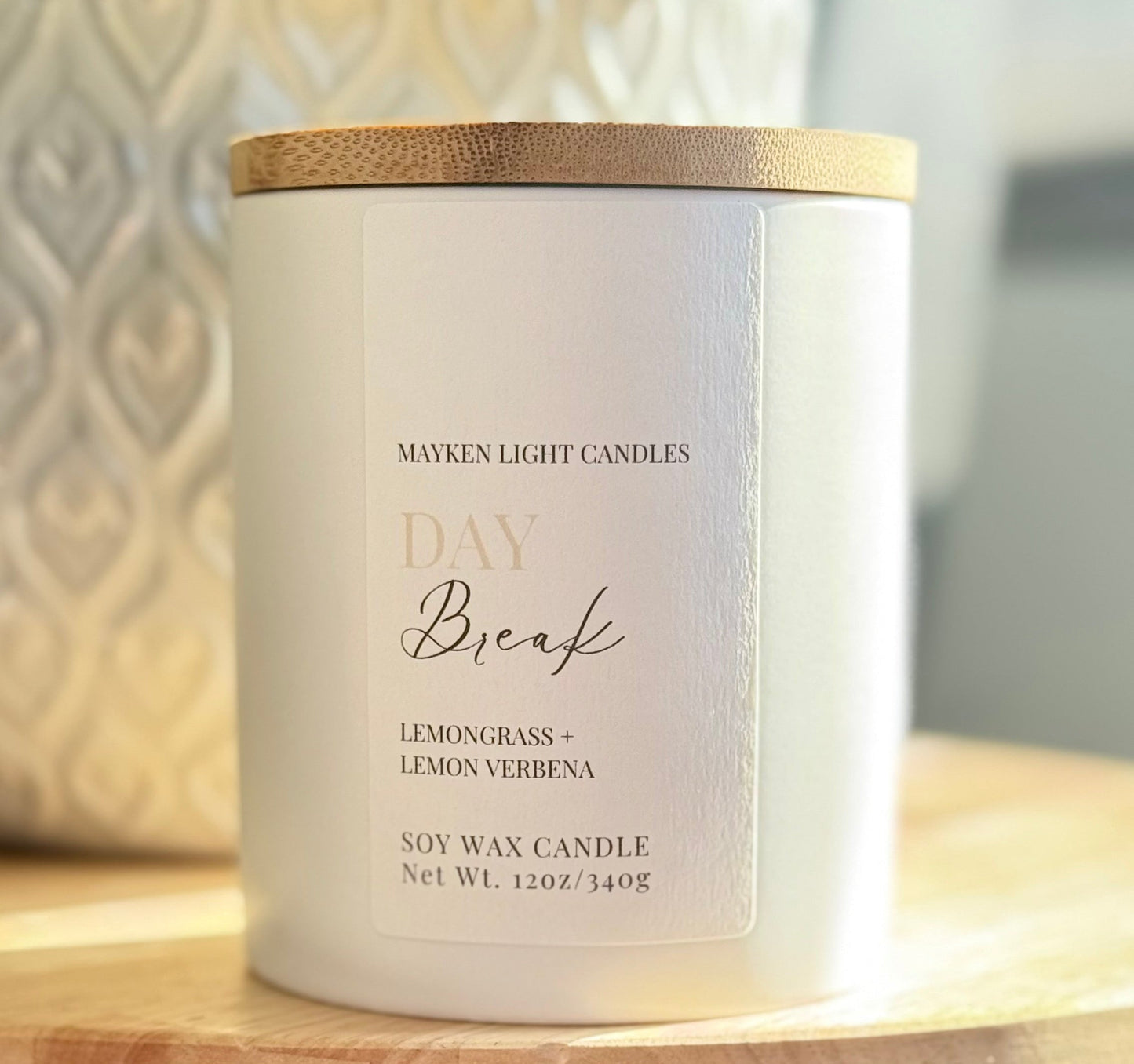 A white candle with a wooden lid, labeled 'DAY BREAK' with the scents 'Lemongrass + Lemon Verbena'. The candle is placed on a wooden surface with a textured background.