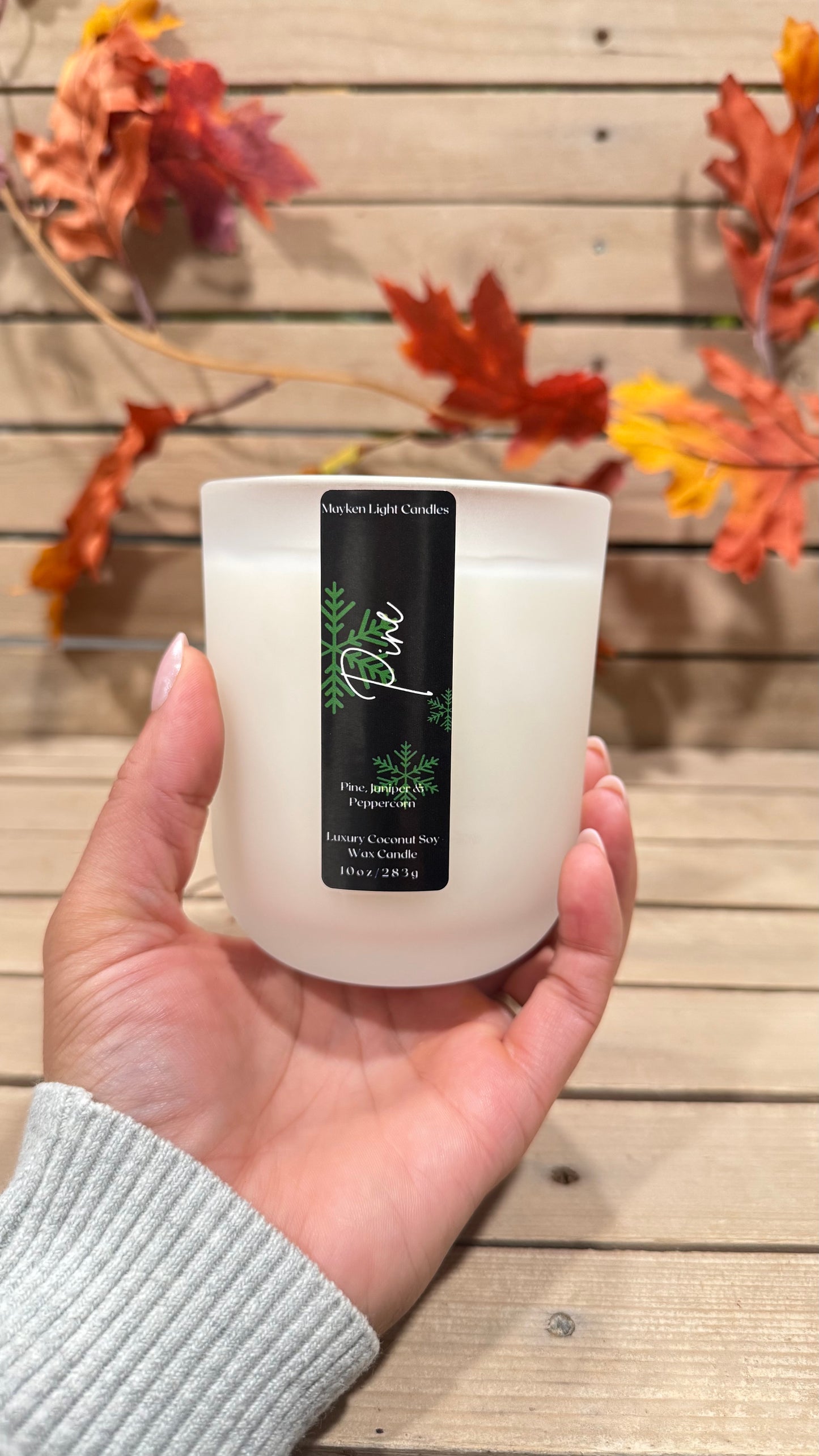 Hand holding a white candle with a label against a wooden background with autumn leaves.