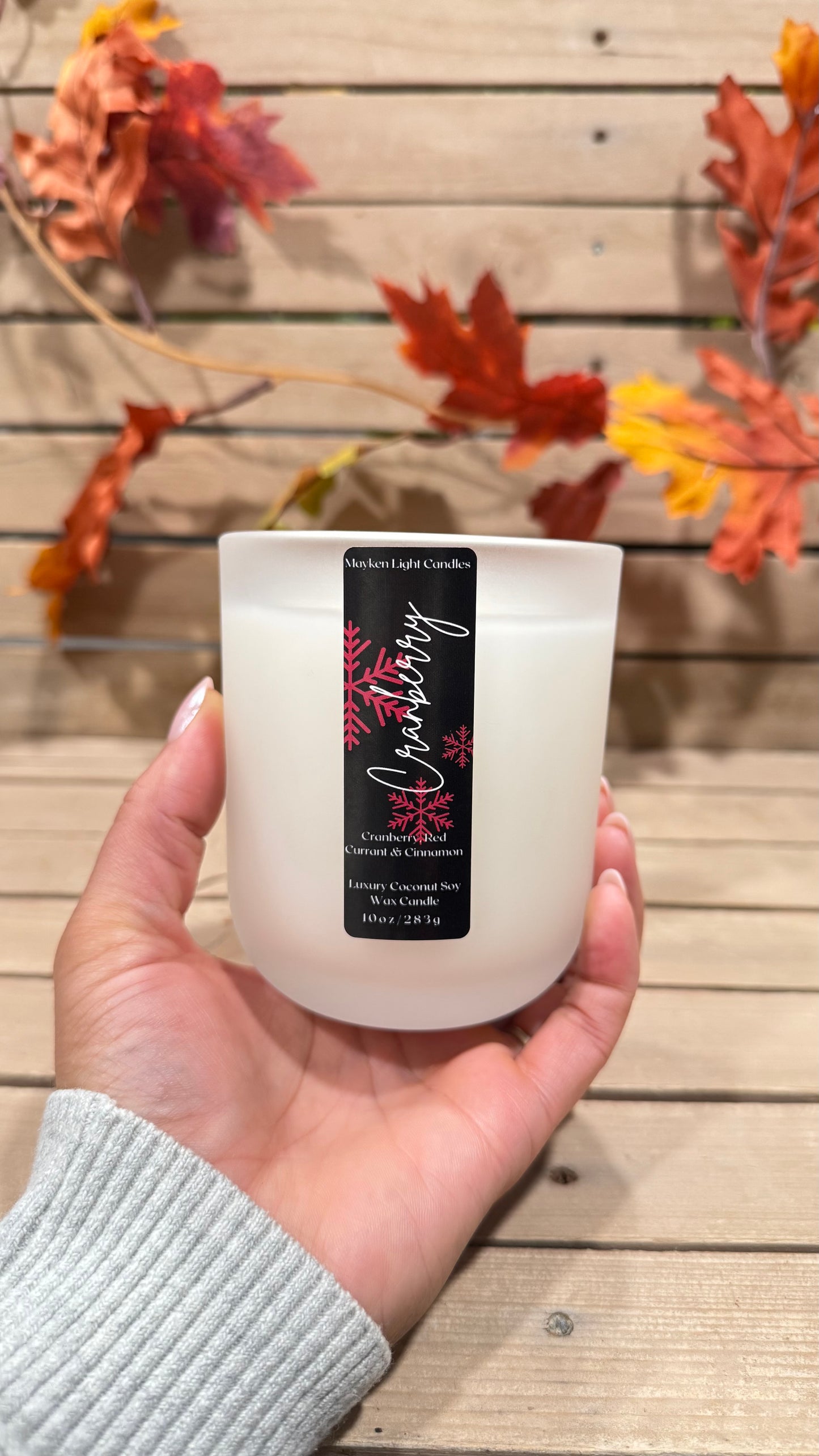 Hand holding a white candle with a black label against a wooden background with autumn leaves.