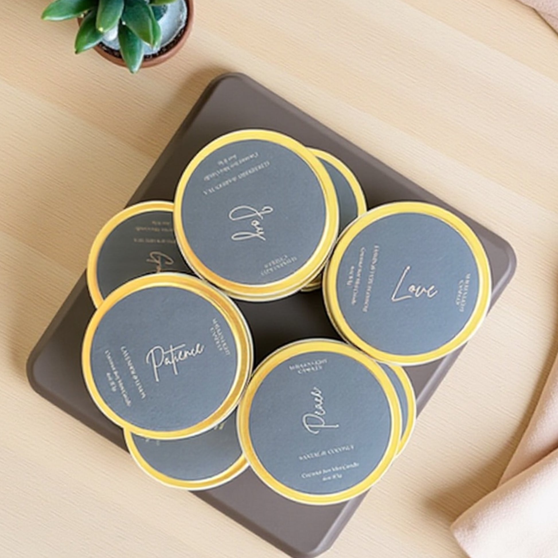 Set of round Gold candle tins with black labels on a dark tray, placed on a light wooden surface.