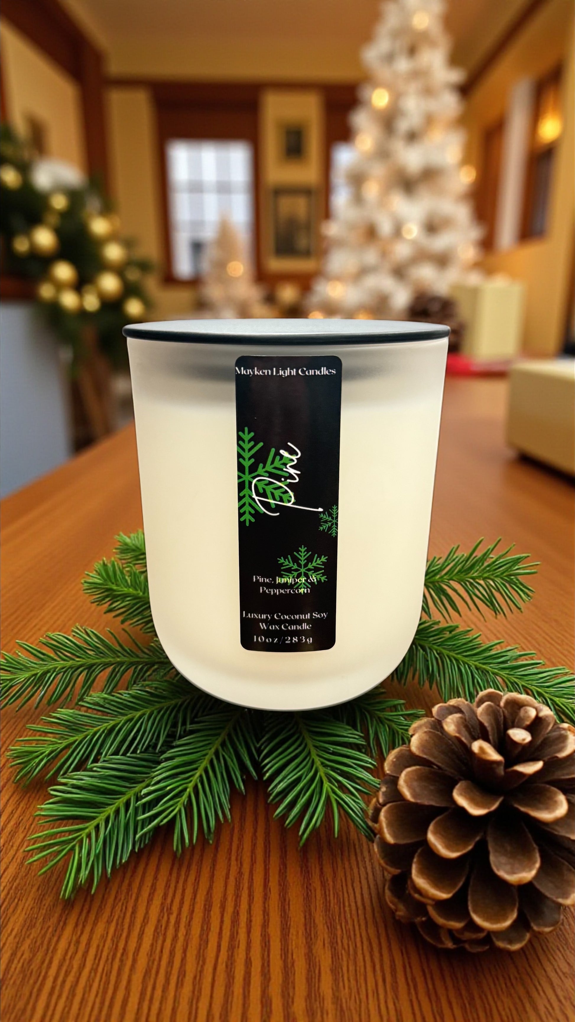 White candle with a black label on a wooden table with greenery and a pine cone.